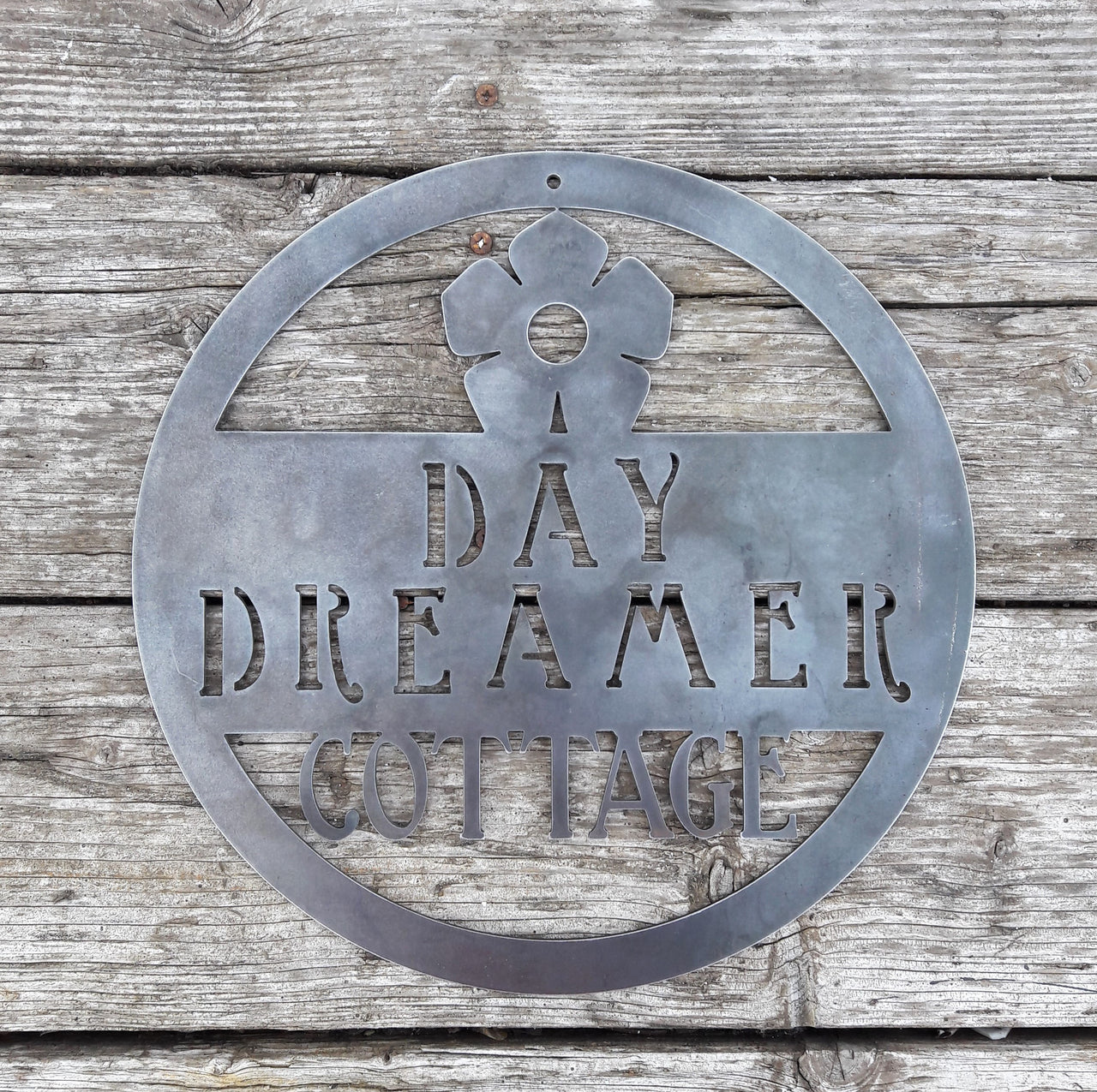 Round metal sign with the image of a flower at the top and three lines of text. The sign reads, " Day Dreamer Cottage".