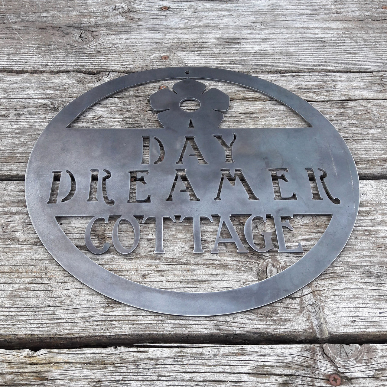 Round metal sign with the image of a flower at the top and three lines of text. The sign reads, " Day Dreamer Cottage".