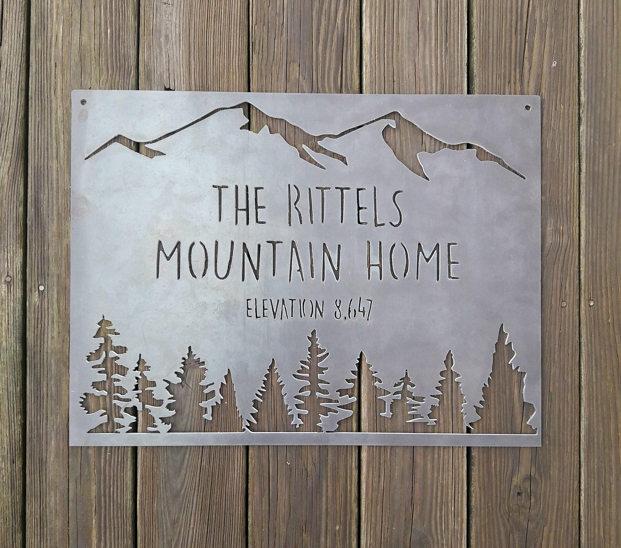 rectangular sign with a mountain range across the top and a coniferous tree line. There are two lines of text on this sign as well as an elevation.  The sign reads, " The Rittels Mountain Home, Elevation 8,647"