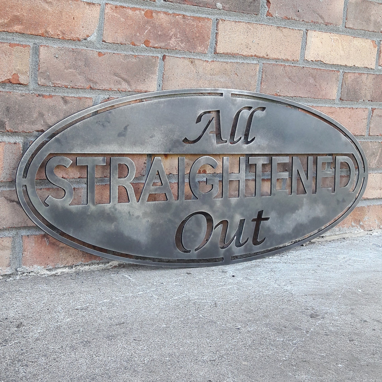 This metal sign is an oval, it has three lines of text. The sign reads, "All Straightened Out"